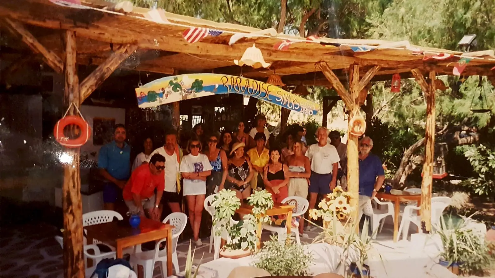 A large group posing under the wooden pergola of Paradise Snacks in the 1990s, below a painted sign