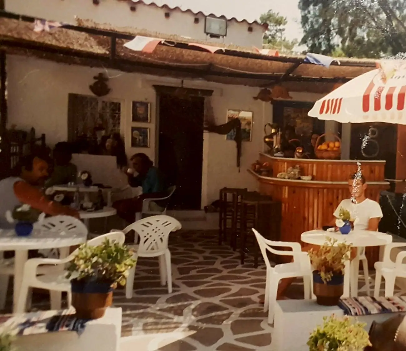The terrace of Snack Bar Paradise in 1995, with a reed roof, a wooden bar, white plastic chairs and a stone floor